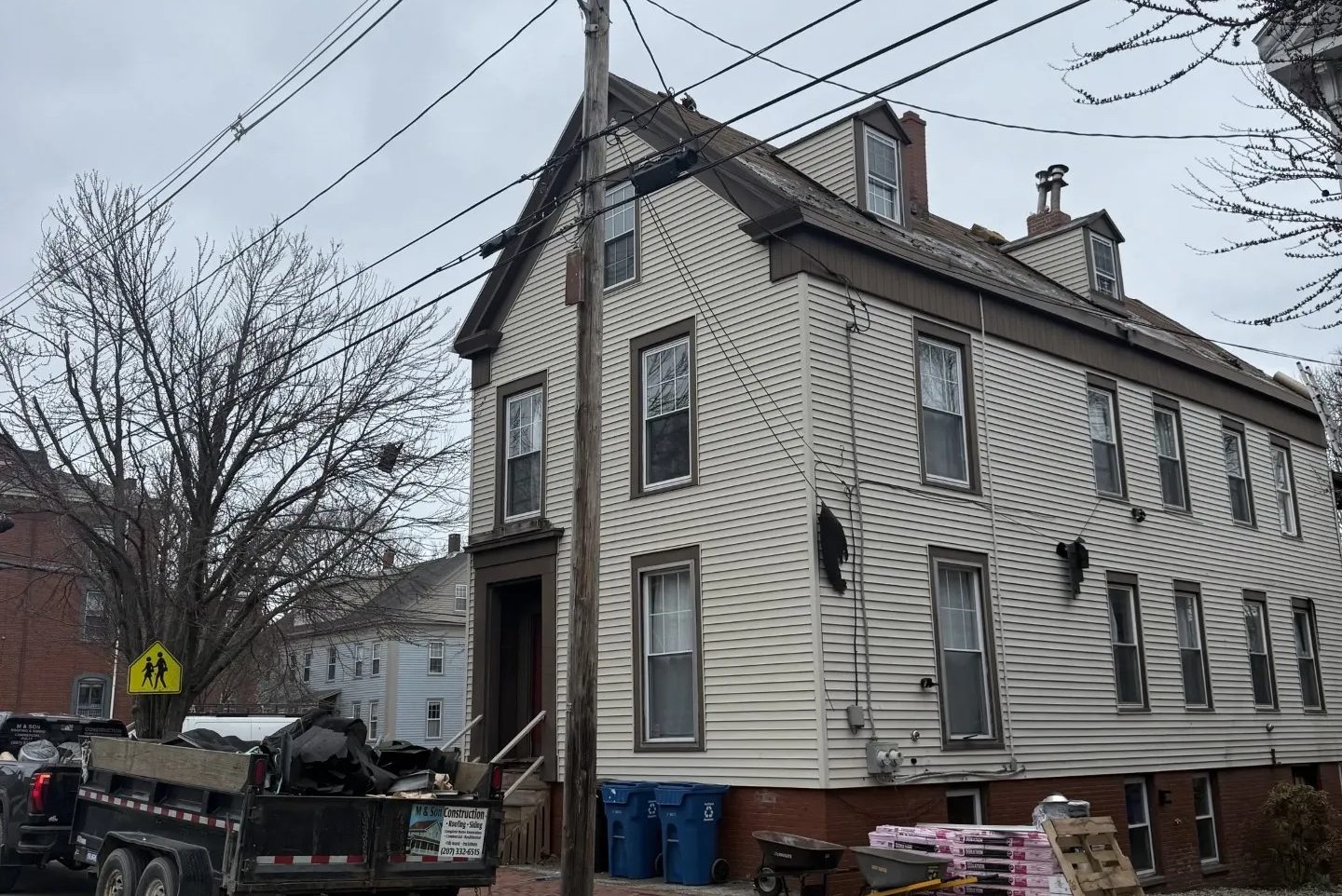 Old white house with peeling siding, construction materials, and parked truck on a snowy street