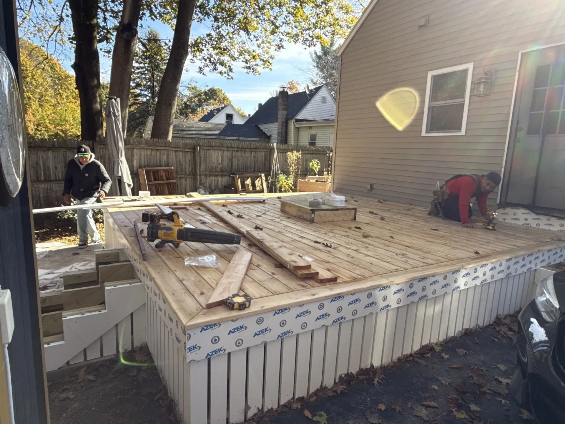 Wooden deck under construction beside a house, with workers, lumber, and tools in the sun.