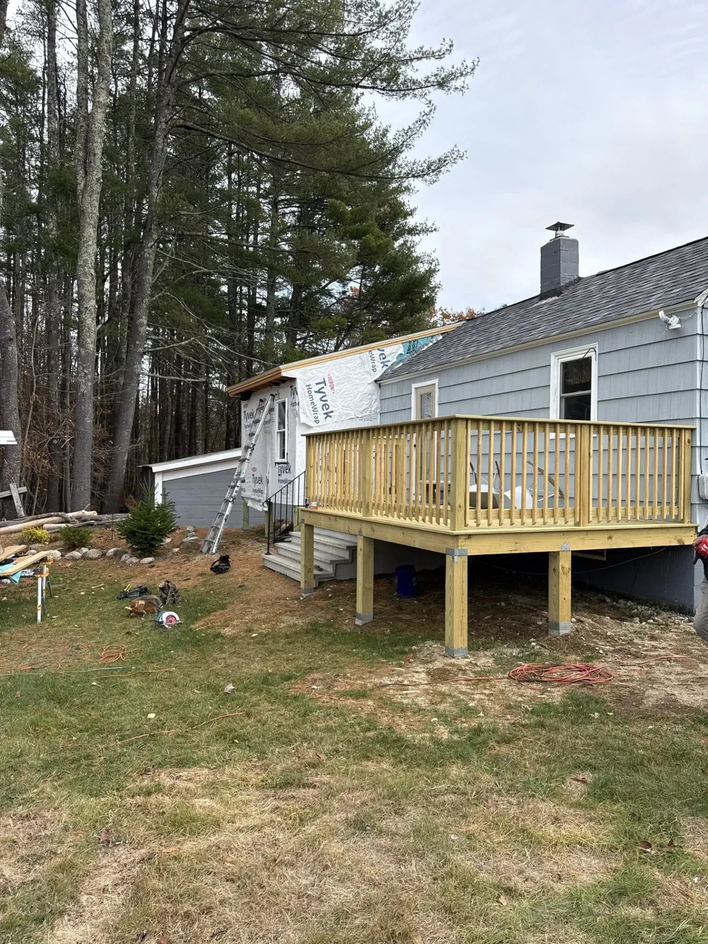 Small gray house with a wooden deck on posts beside trees and a grassy yard