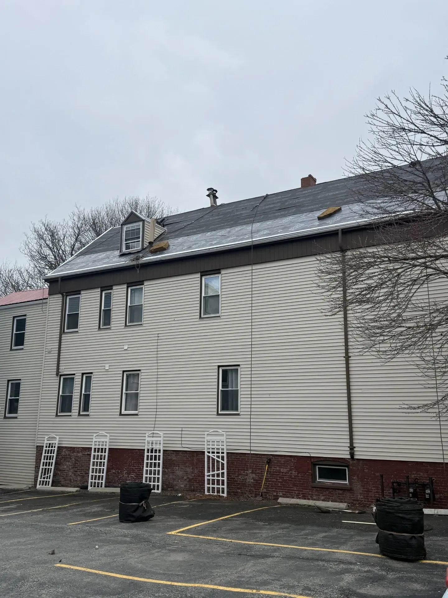 Three-story beige apartment building with white trim, red brick foundation, and parking lot in front