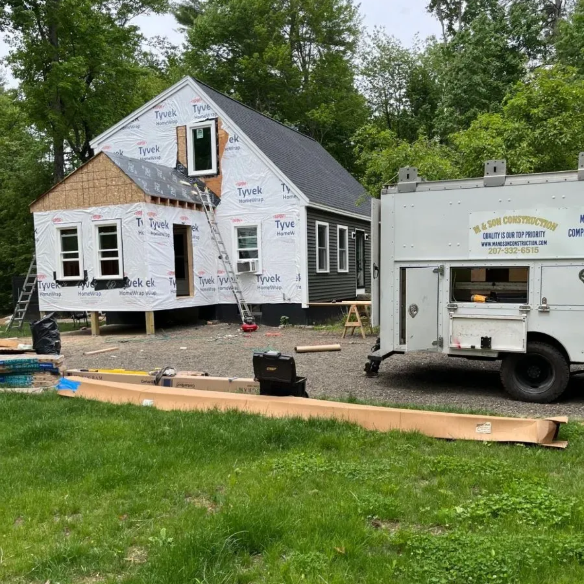 House under construction with white wrap, ladder, and utility trailer in a grassy yard