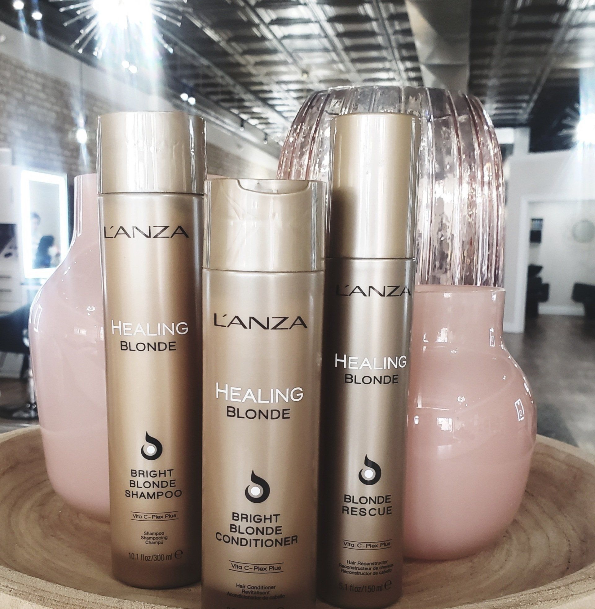 Three L'anza Healing Blonde hair products in gold bottles with two pink vases in a salon setting.