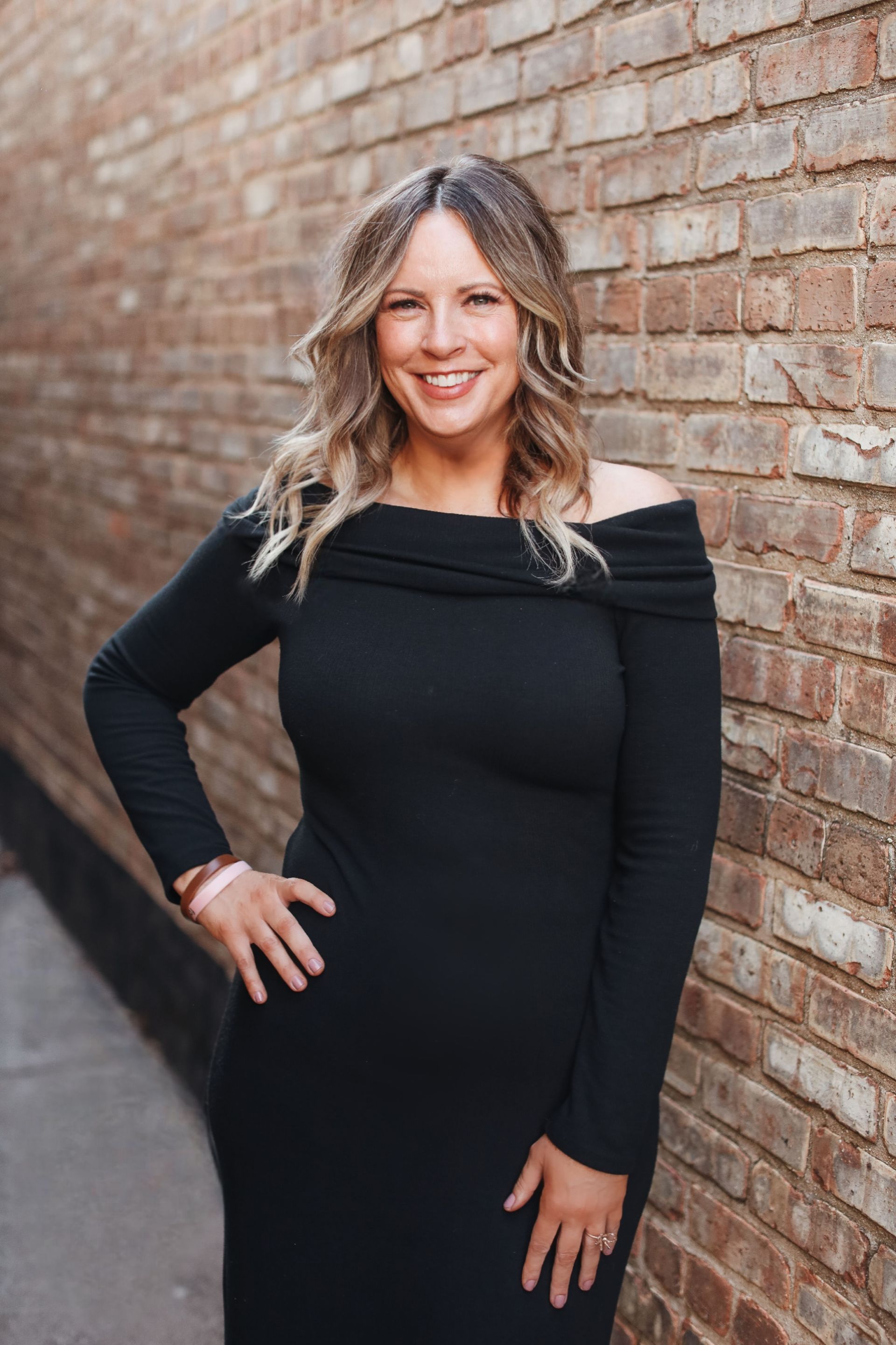 Woman in black top smiles, leaning against a brick wall.