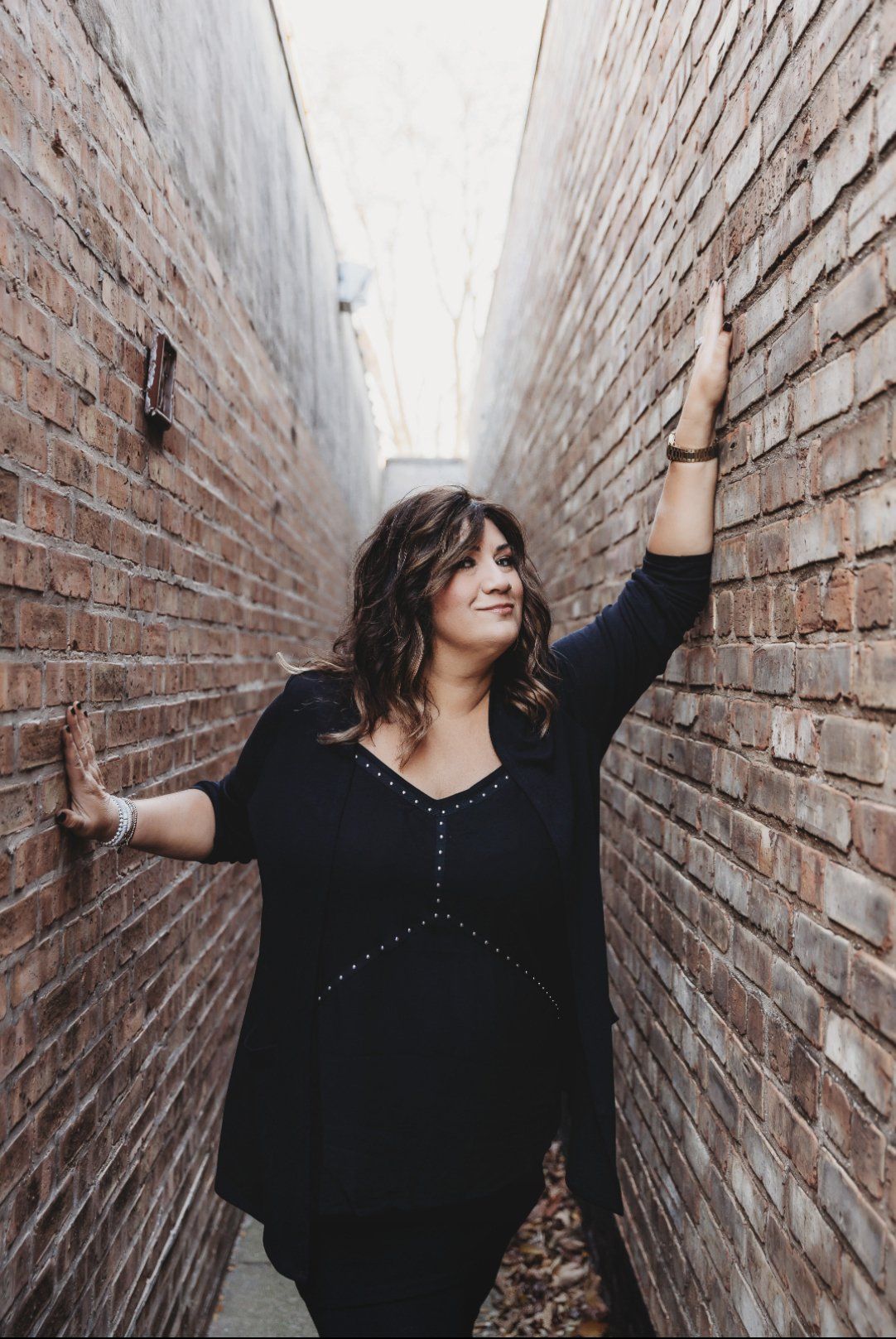 Woman in black sweater leans against brick walls in alley, smiling.