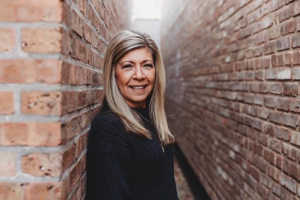 Woman with blonde hair smiles, leaning against a brick wall in a narrow alley.