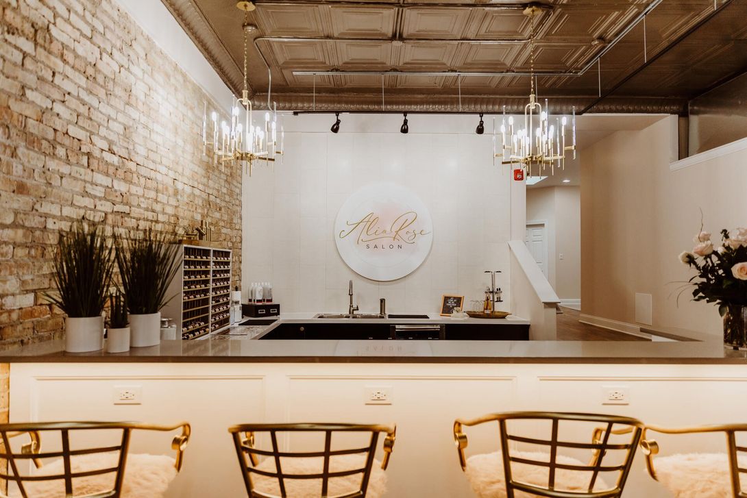 Nail salon interior with exposed brick wall, bar with seating, and decorative lighting.