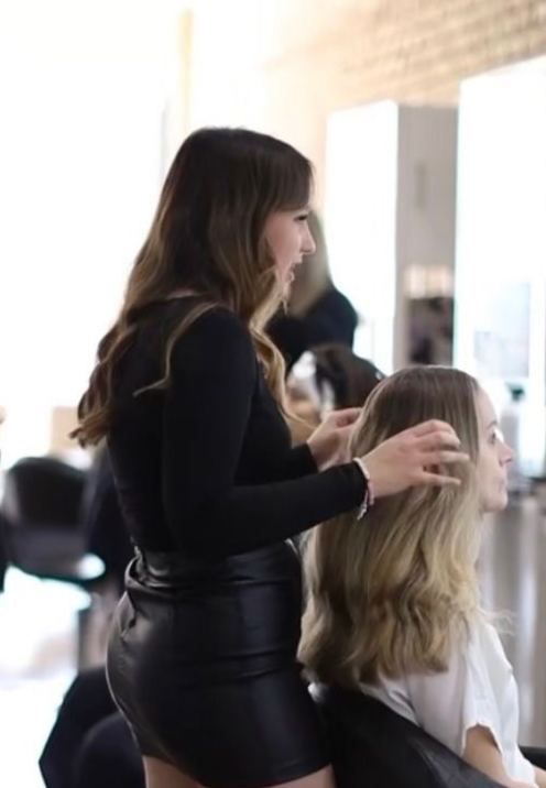 A hairstylist working on a client's hair in a salon. Both are light-skinned; stylist has brown hair and client has blonde.