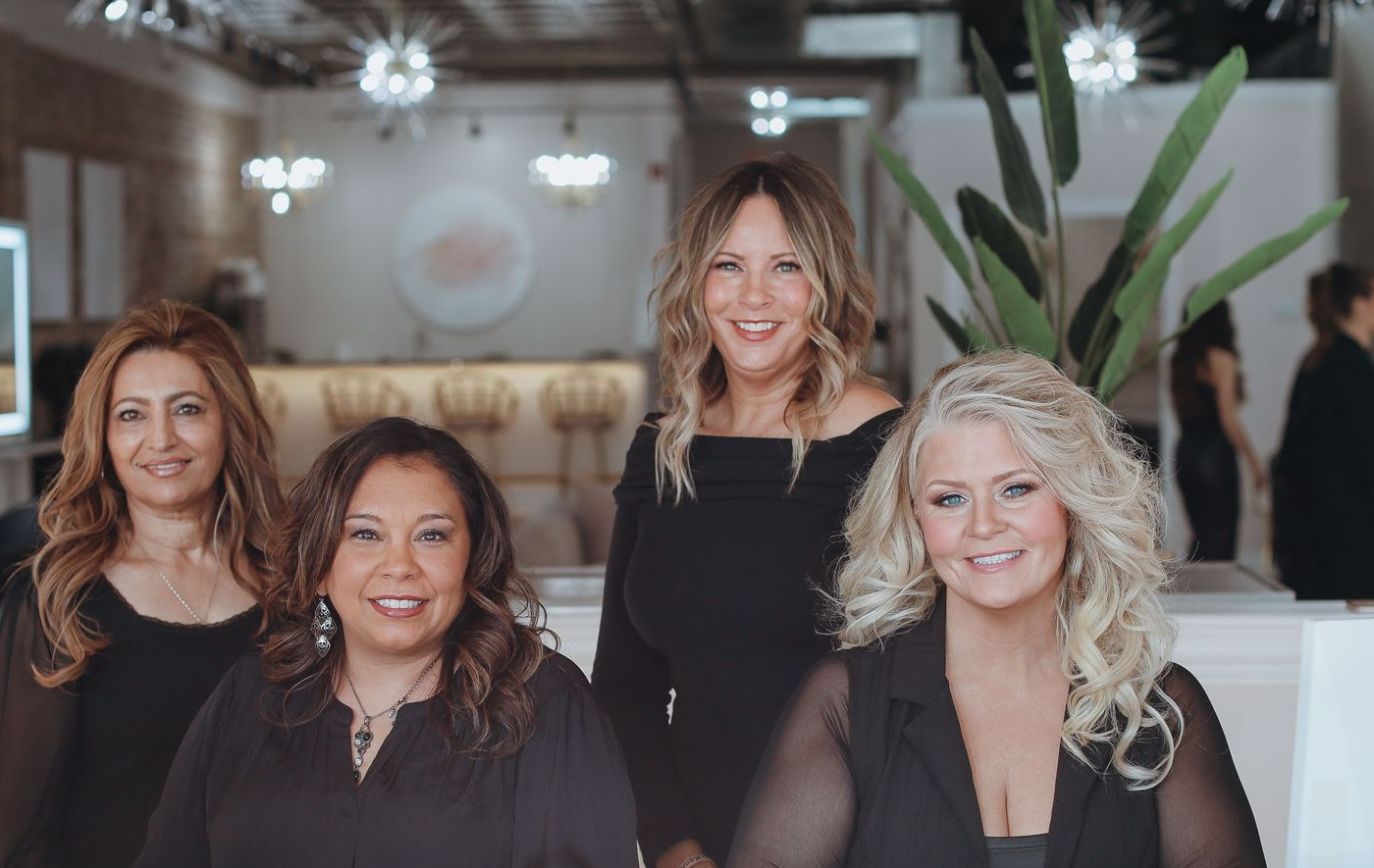 Four women smiling in a well-lit salon setting with plants and decor.