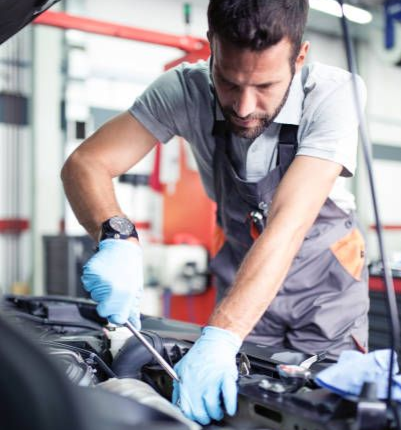 Professional automotive technician leaning over a car engine bay performing focused repair work.