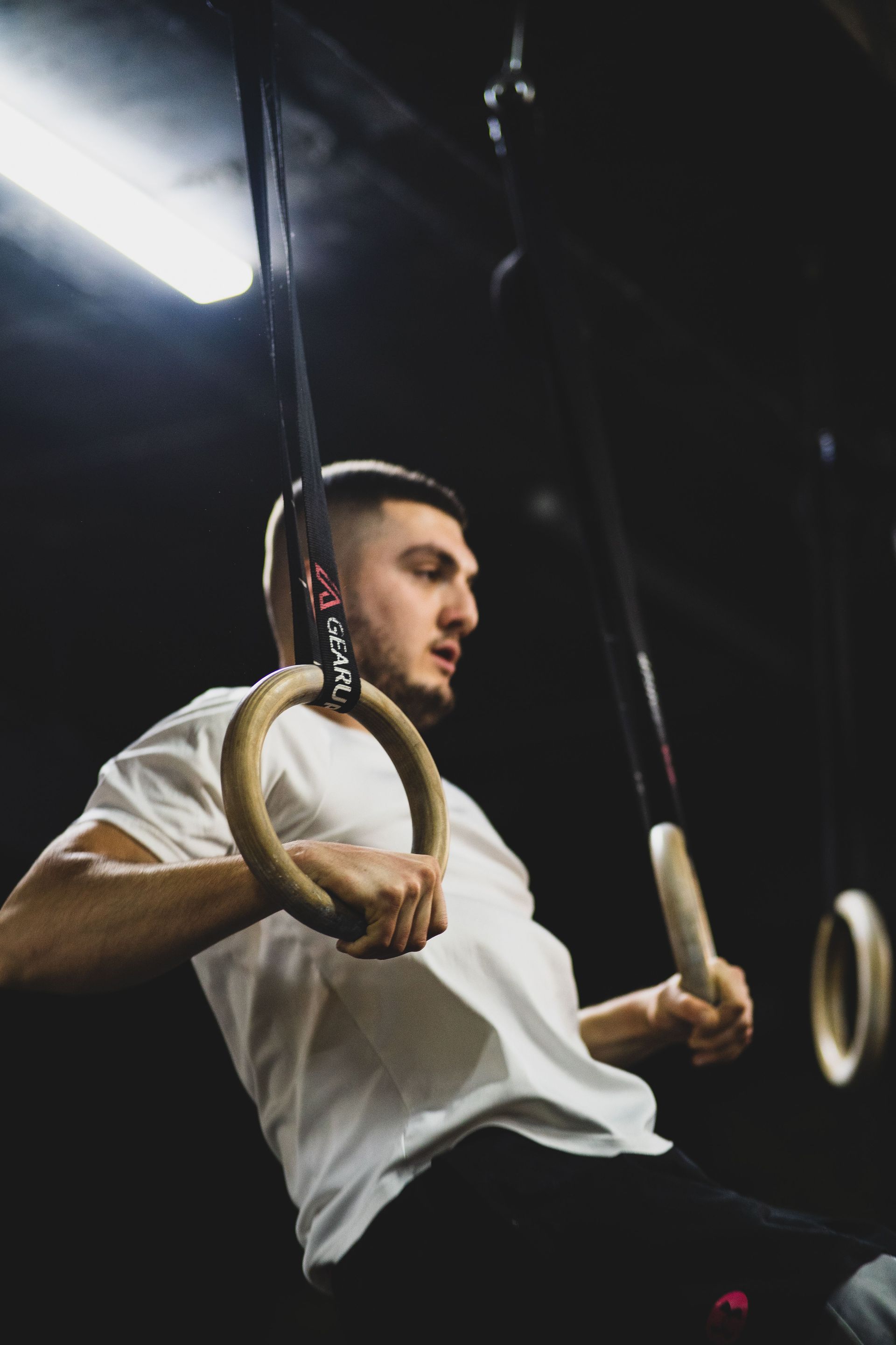 Man performing ring dips on gymnastic rings in a dark gym, wearing a white shirt.