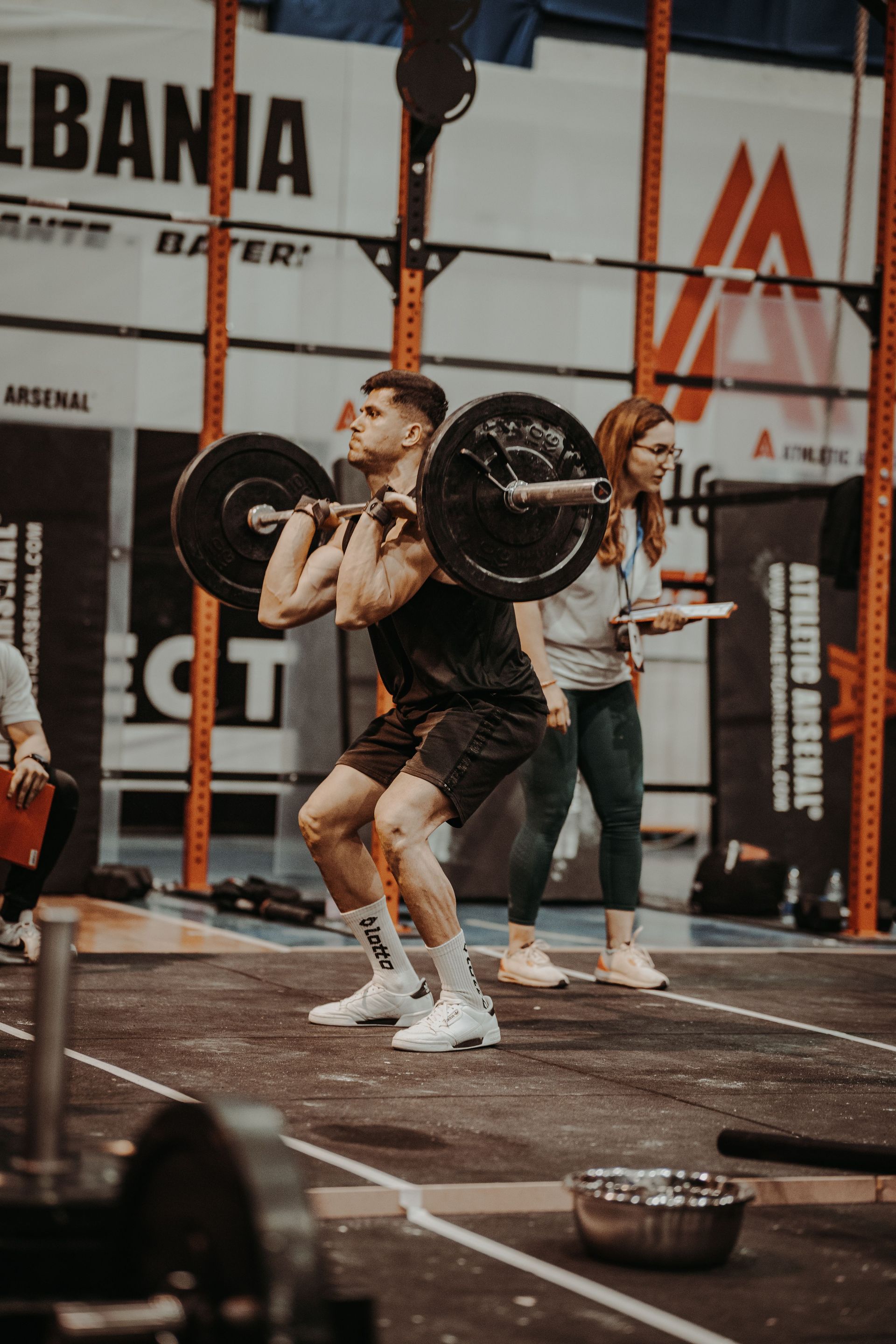 Athlete lifting a barbell in a gym, with a trainer behind him and equipment in the background