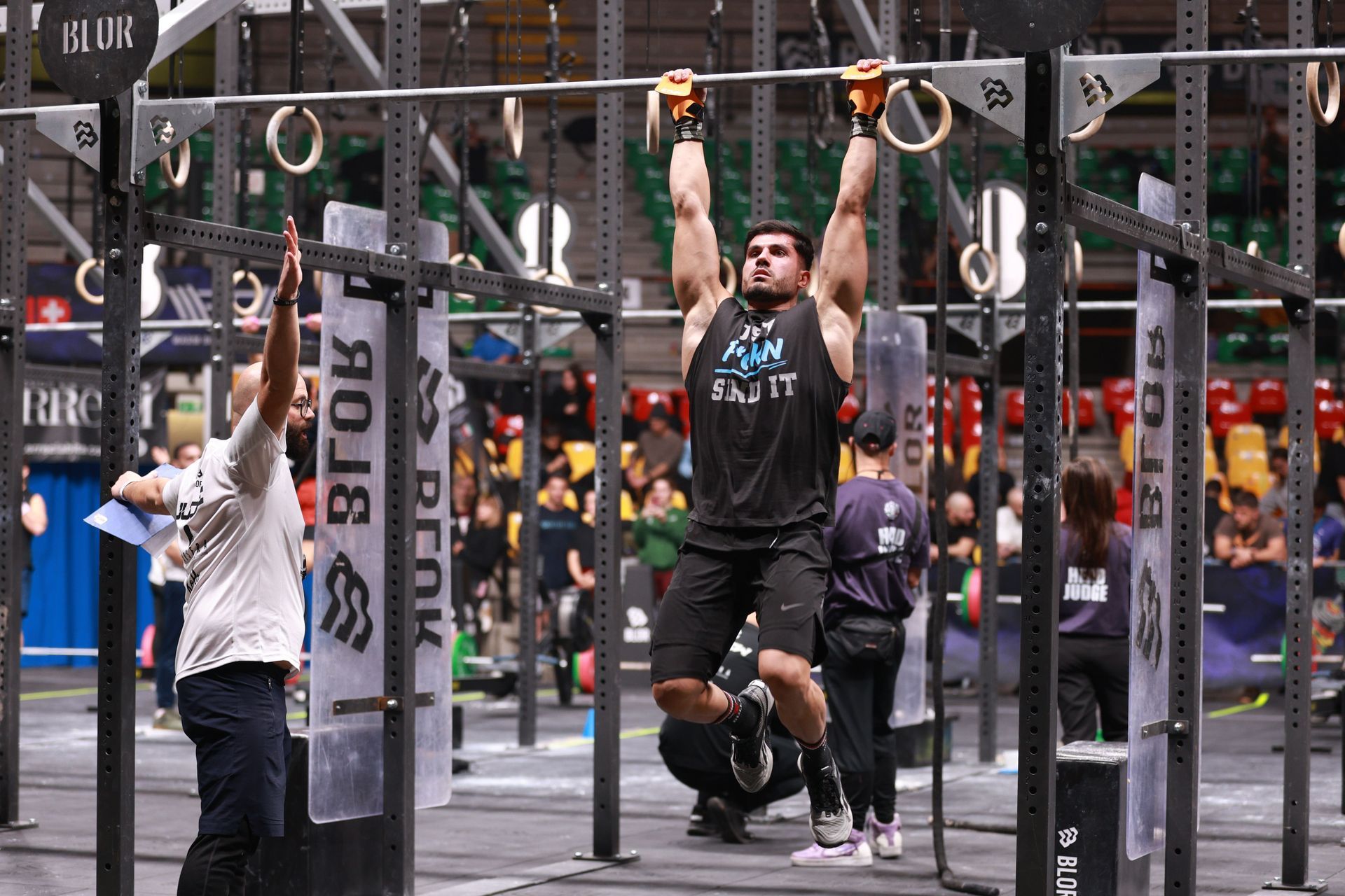 Athlete doing pull-ups on rings in an indoor competition, with judges and spectators nearby