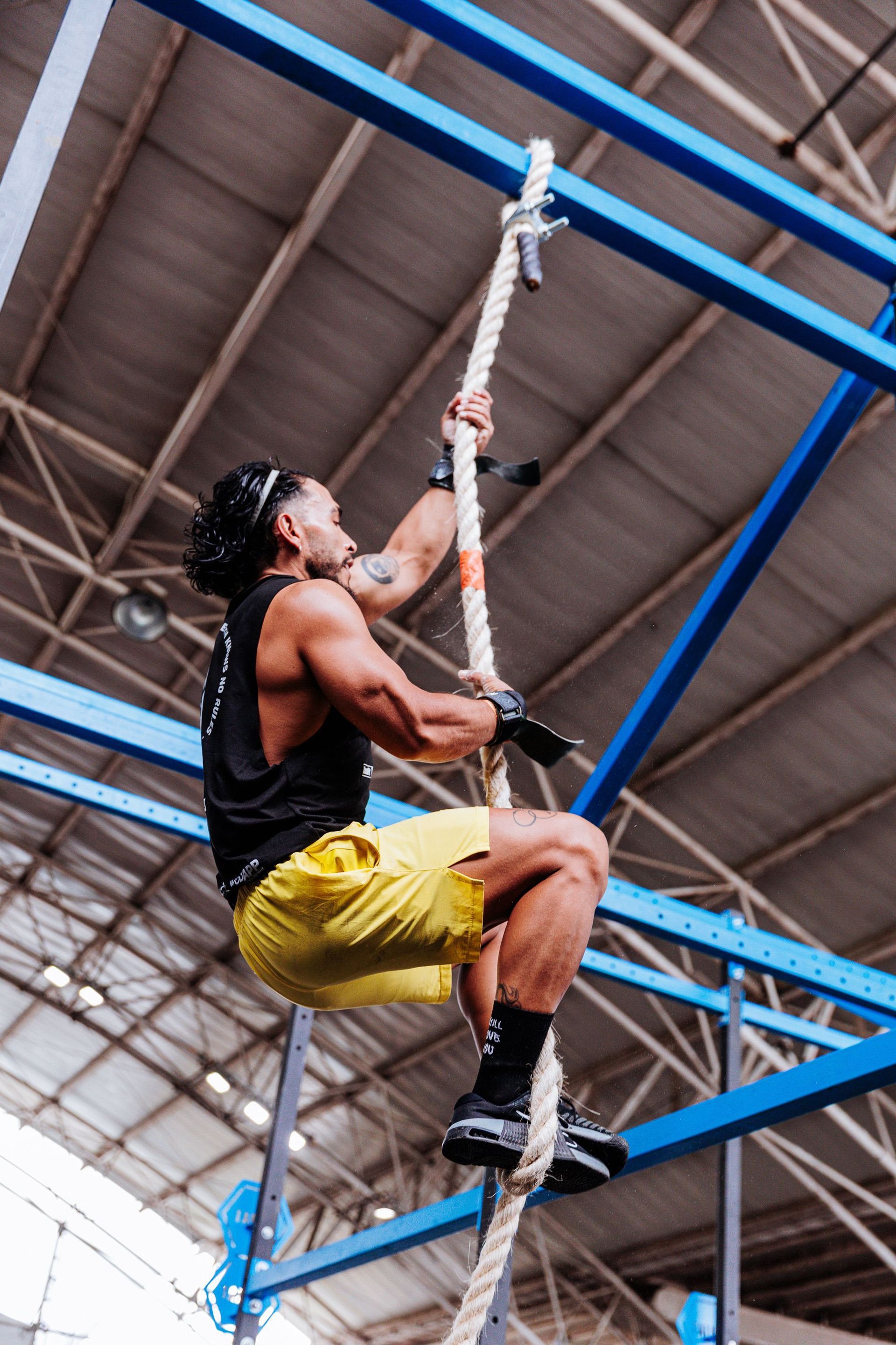 Man climbing a rope indoors in a gym, wearing black tank top and yellow shorts.