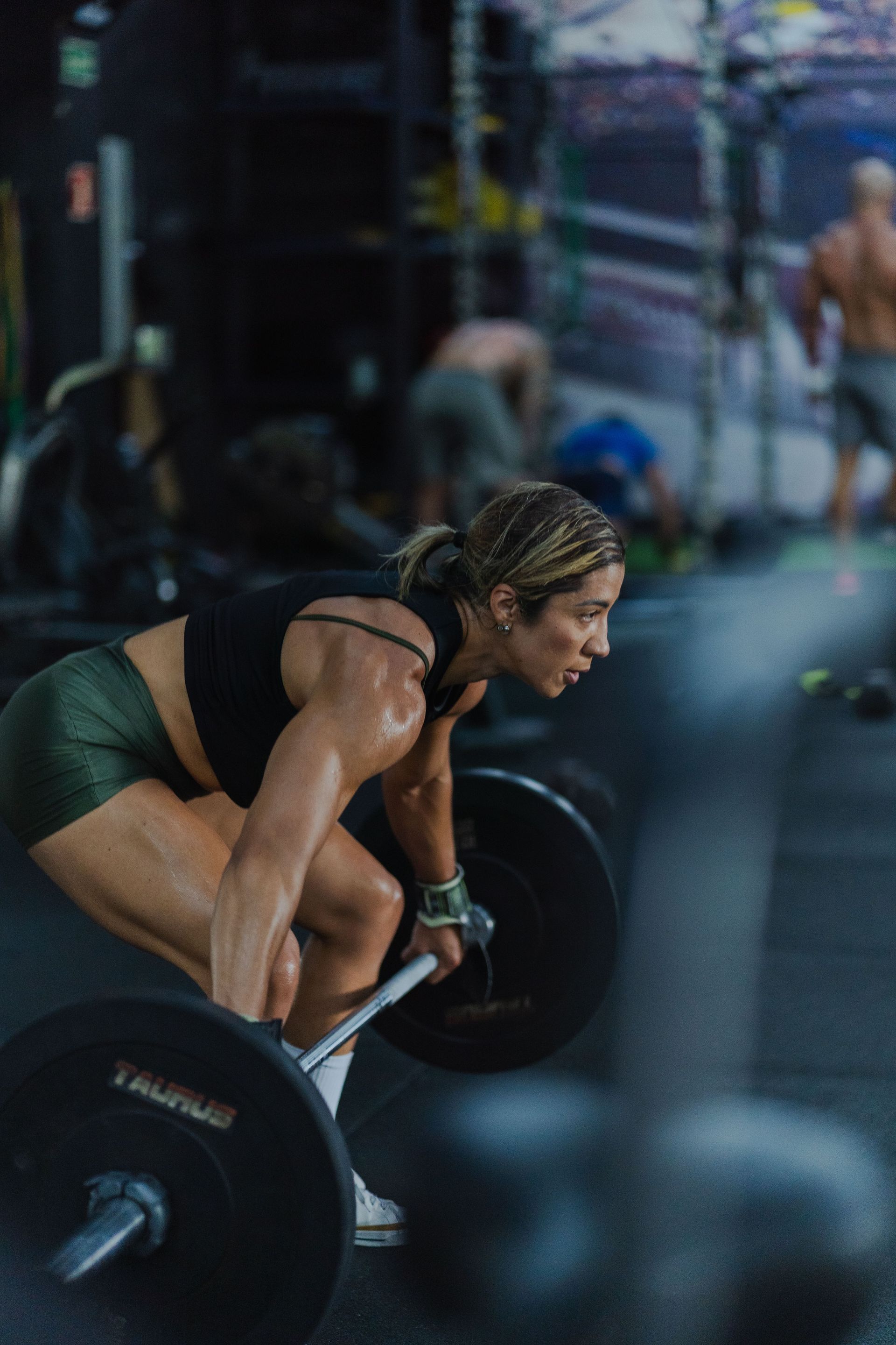 Woman lifting a barbell in a gym, bent over in a deadlift position with focused expression.