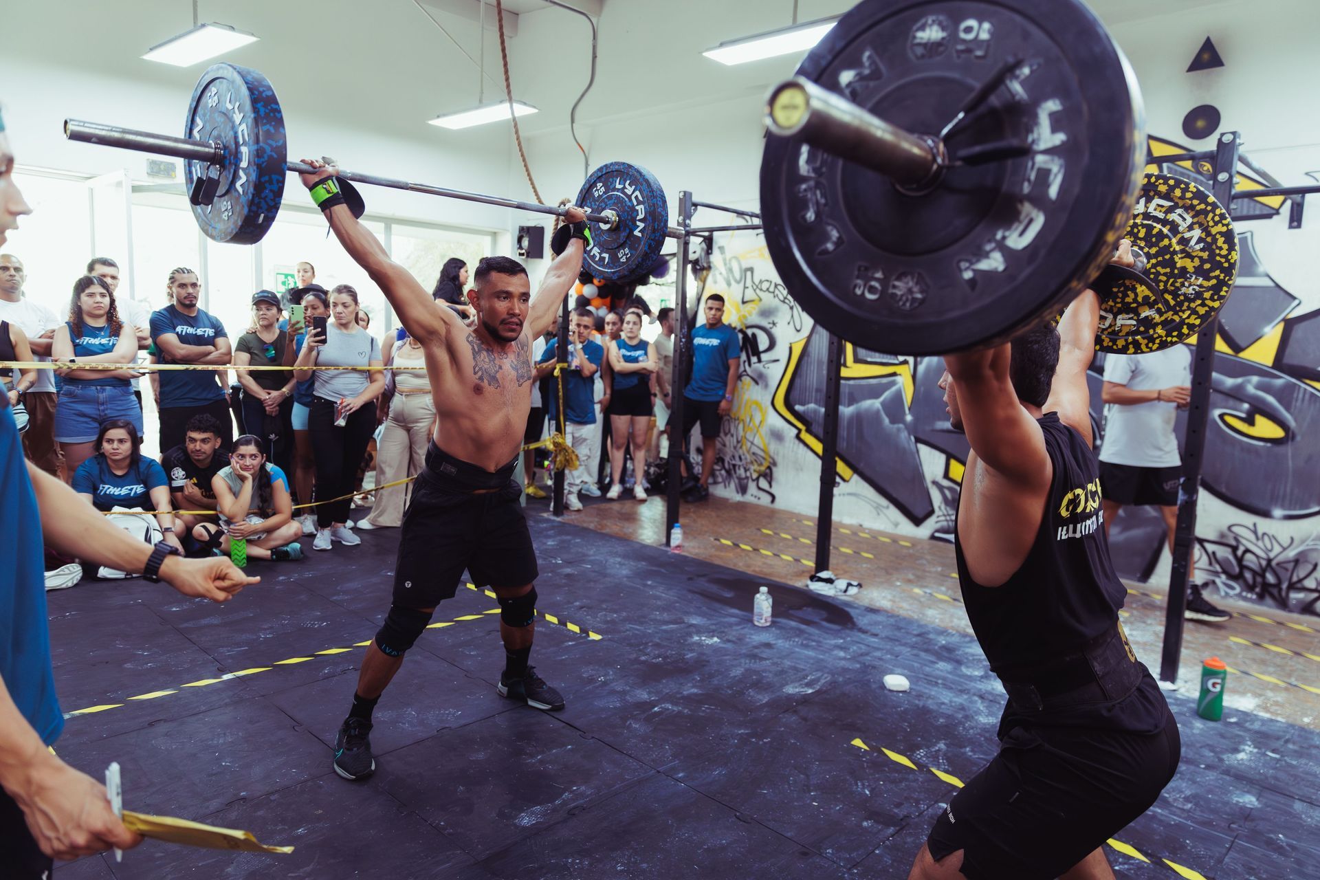 Two athletes lifting barbells overhead in a crowded gym while spectators watch