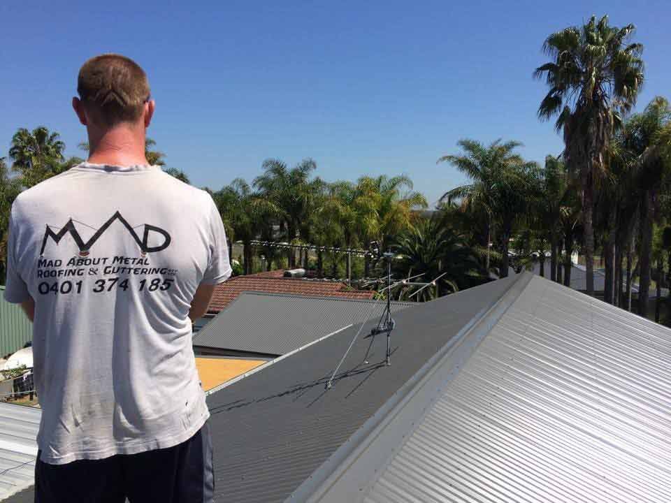 A Man Wearing A Mad Shirt Is Standing On A Roof — Mad About Metal Roofing & Guttering PTY LTD in Newcastle, NSW