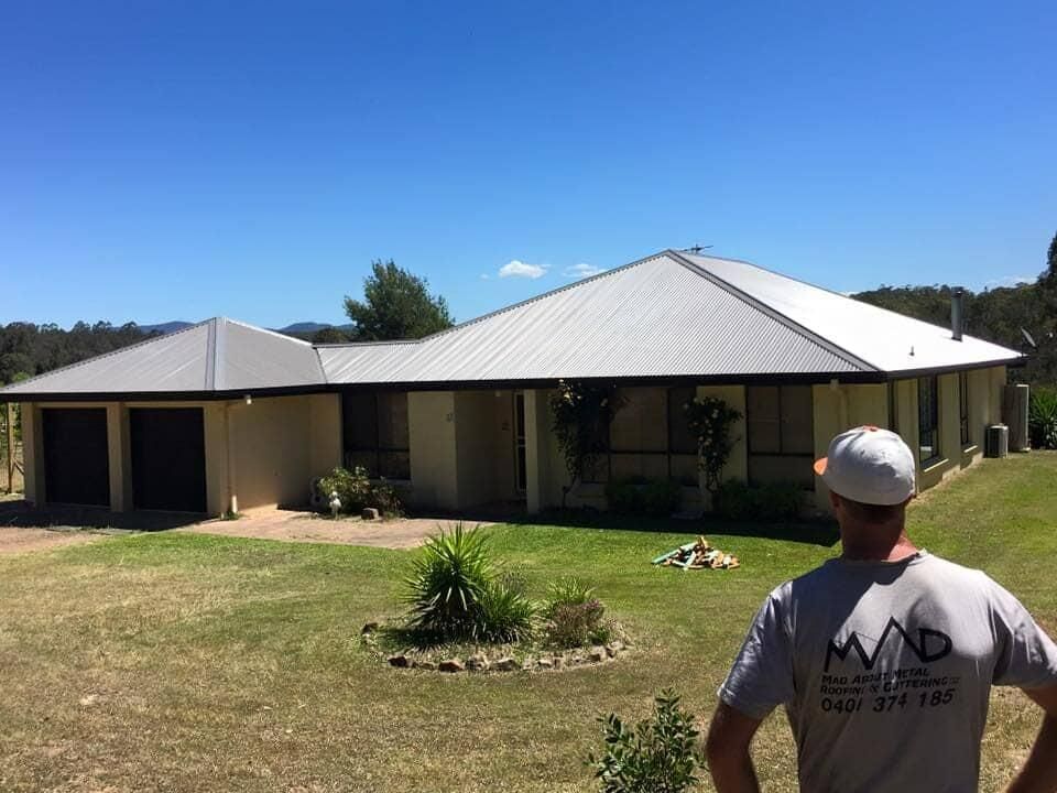 A Man Is Standing In Front Of A Large House With A White Roof — Mad About Metal Roofing & Guttering PTY LTD in Newcastle, NSW