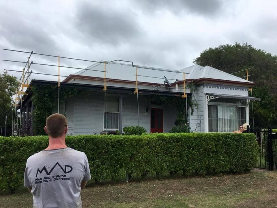A Man Is Standing In Front Of A House With Scaffolding On The Roof — Mad About Metal Roofing & Guttering PTY LTD in Newcastle, NSW