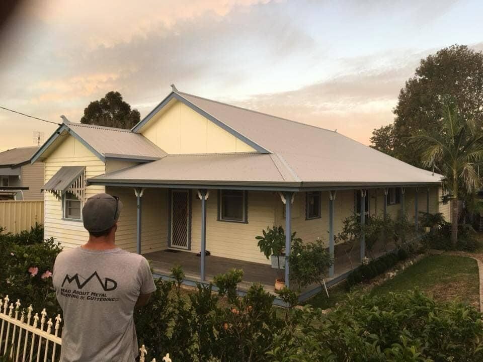 A Man In A Mad Shirt Stands In Front Of A House With New Roof — Mad About Metal Roofing & Guttering PTY LTD in Newcastle, NSW