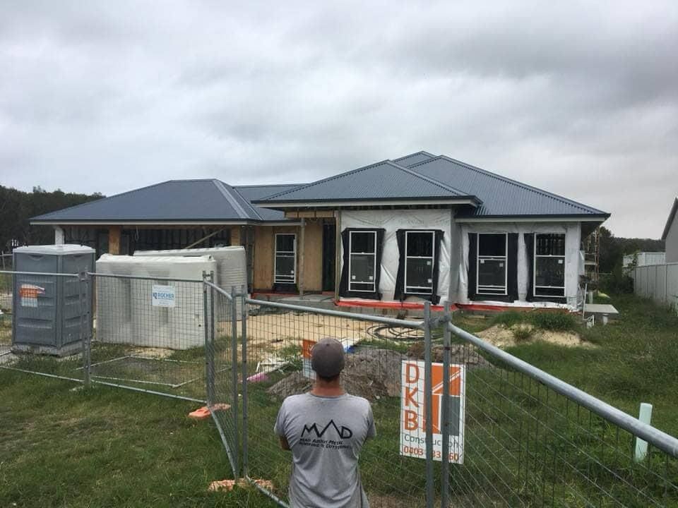 A Man Is Standing In Front Of A House Under Construction Behind A Fence — Mad About Metal Roofing & Guttering PTY LTD in Newcastle, NSW