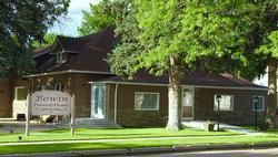 Brick building with a sign, large windows, and green trees in front on a sunny street.