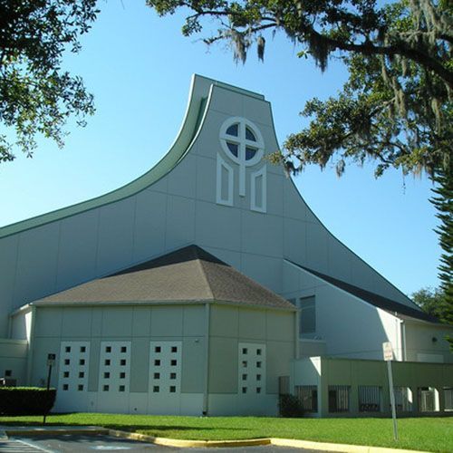 Modern church with a curved gray roof and cross window, framed by trees under a blue sky