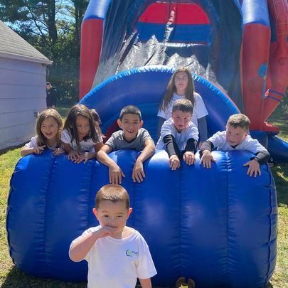 Seven children pose in front of a blue inflatable slide on a sunny day. One child is in front, six lean over the barrier.