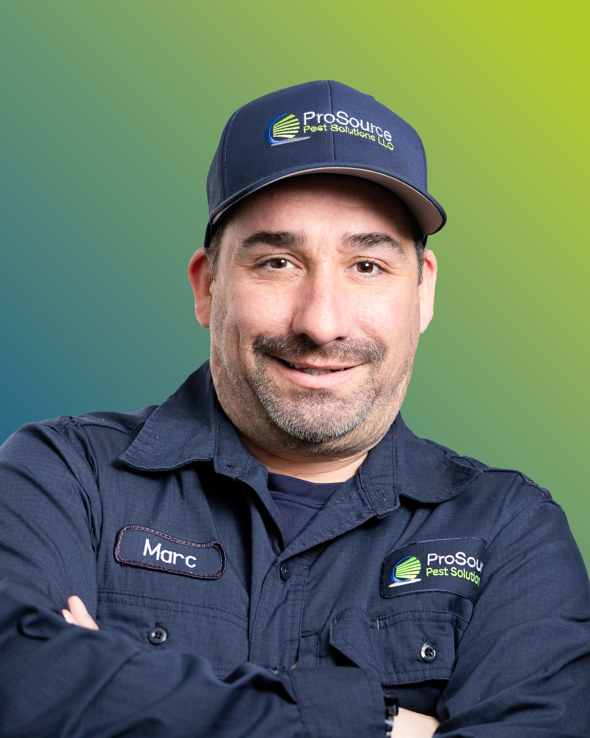 A smiling person wearing a dark blue button-down shirt with a name tag and a branded cap against a green background.