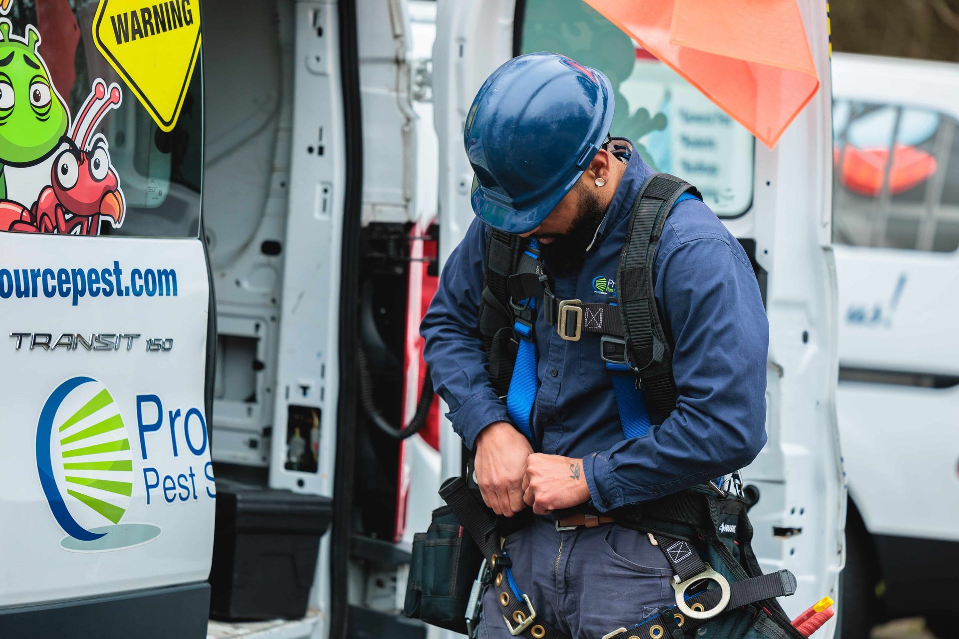 A pest control worker wearing a blue hard hat and safety harness stands beside a branded company van.