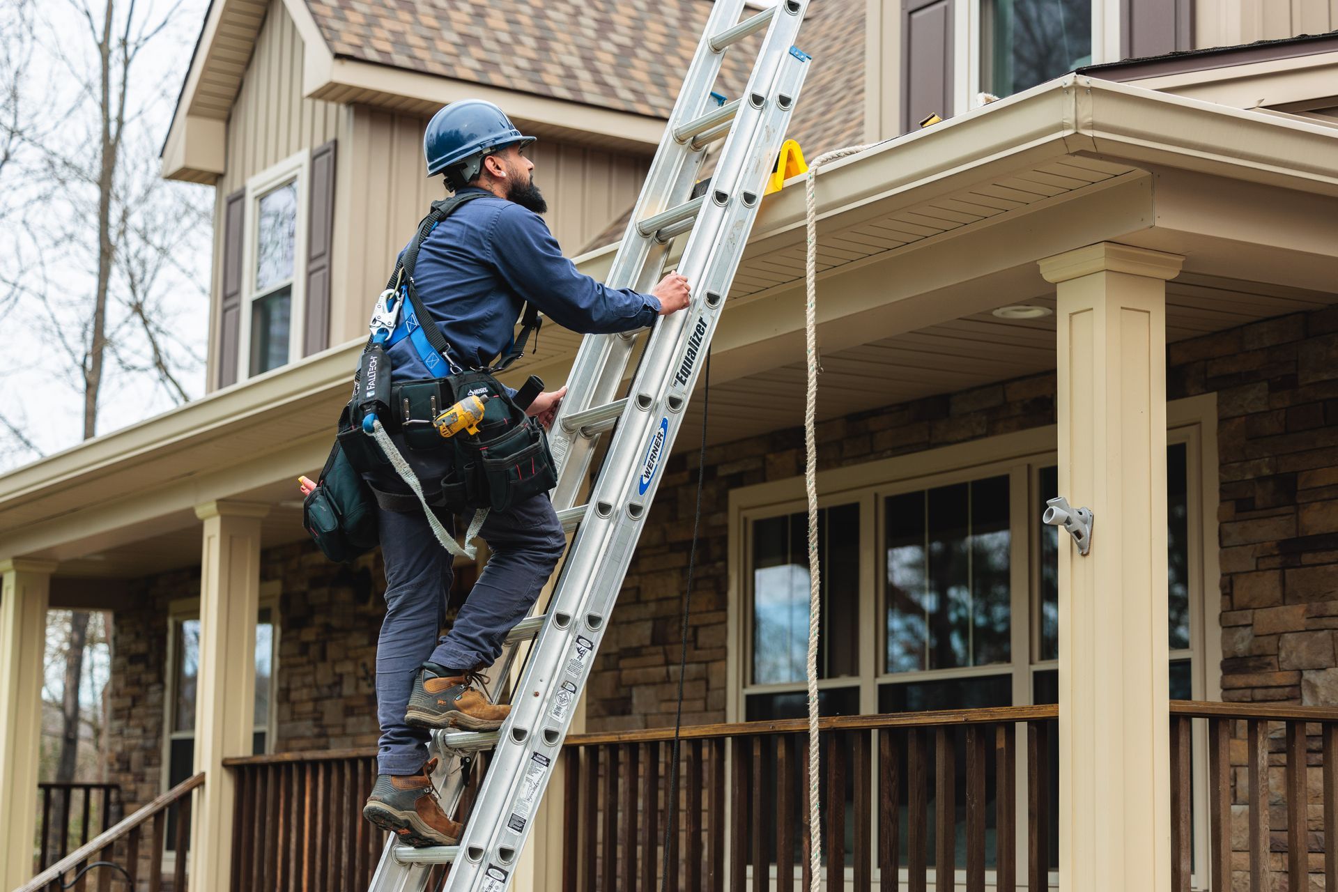 A technician in a hard hat and safety harness climbs an aluminum ladder positioned against the side of a suburban home.