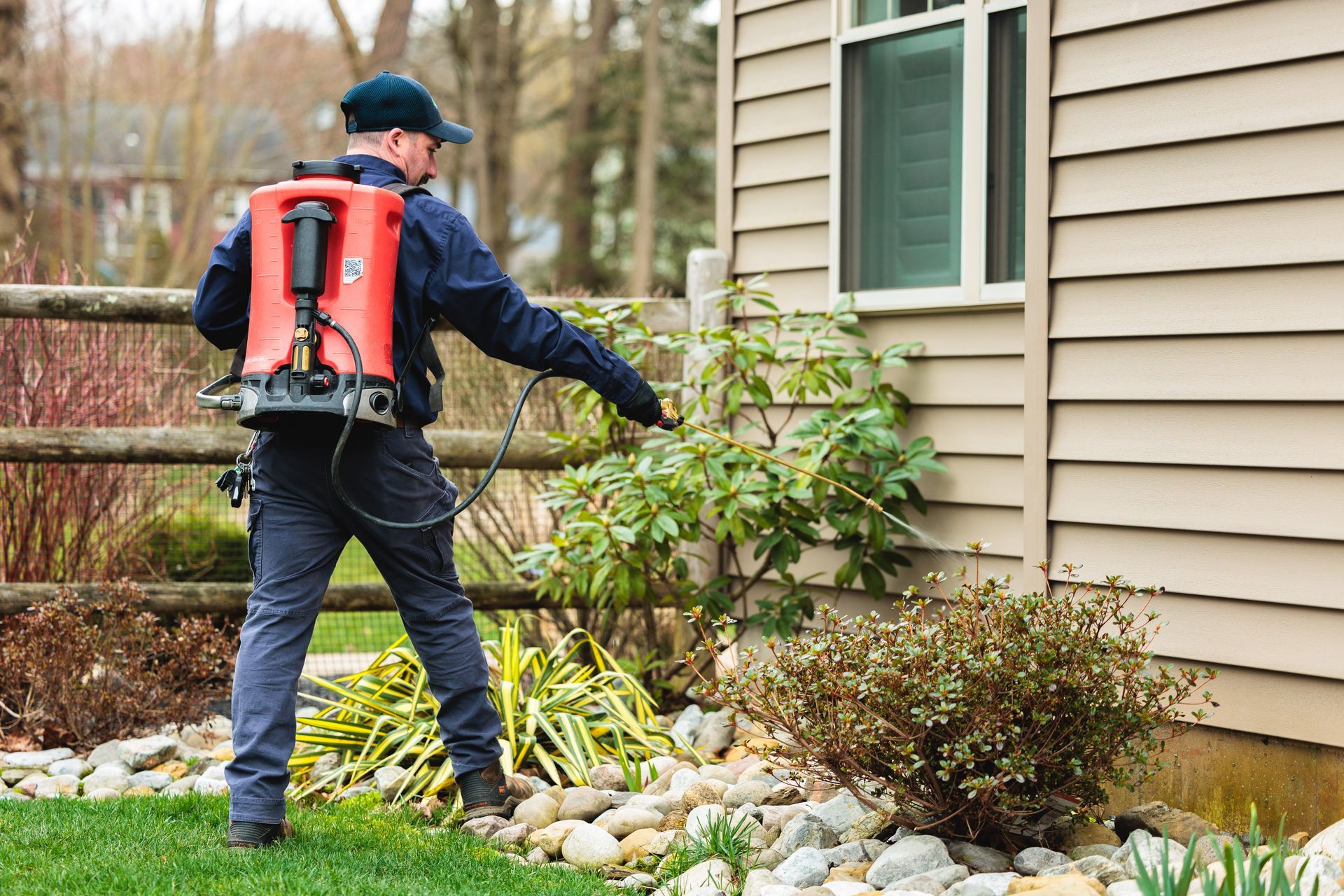 A person in work uniform wearing a red backpack sprayer treats the exterior of a house with pest control spray.