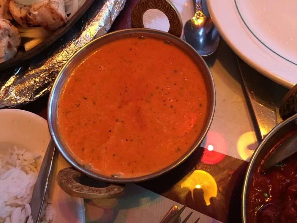 A bowl of orange curry sauce on a restaurant table, surrounded by other dishes and silverware.