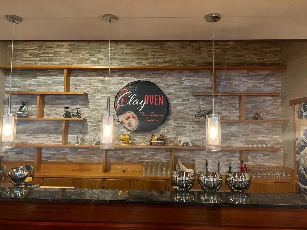 Restaurant bar area with wooden shelves holding small objects; stone-like wall, pendant lights, and large logo.
