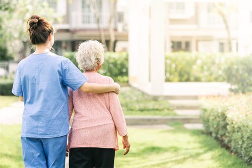 A caregiver in blue scrubs walks outdoors with an arm around an individual in a pink shirt, providing support.