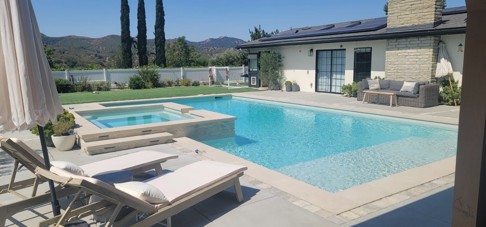 A sunny outdoor patio with a swimming pool and attached hot tub, featuring lounge chairs under an umbrella and a house.
