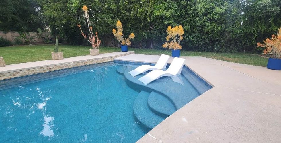 Two white lounge chairs partially submerged in the shallow sun shelf of a blue swimming pool in a garden setting.