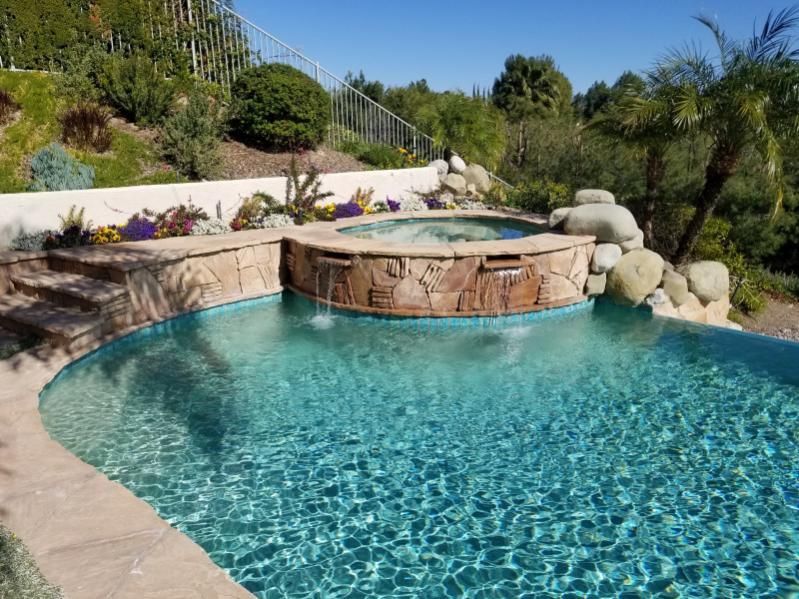 An outdoor swimming pool with a raised stone hot tub featuring two cascading water spouts, set in a landscaped backyard.