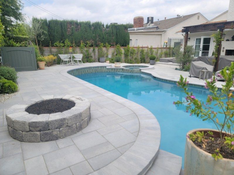 A backyard pool area featuring a curved stone fire pit on a light grey paver patio next to a swimming pool and hot tub.