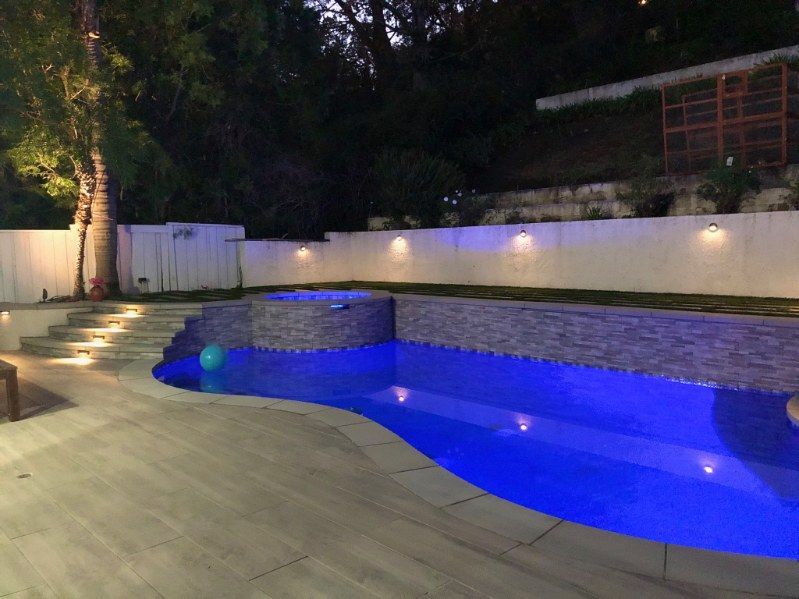A backyard swimming pool with blue lighting, a stone-tiled wall, and steps illuminated at night.