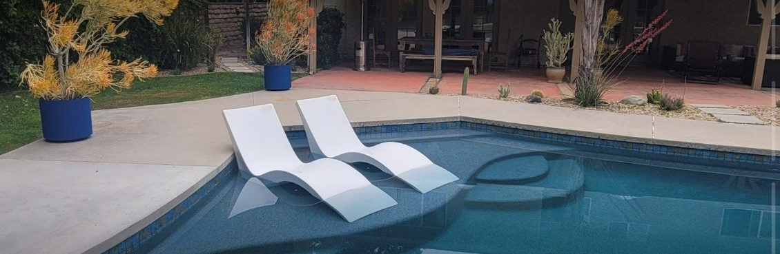 Two white lounge chairs sit in the shallow water of a swimming pool on a patio with potted plants in the background.