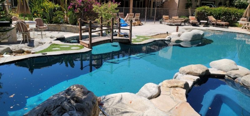 A backyard pool area featuring a wooden bridge, rock accents, a built-in spa, and lounge chairs on a sunny patio.