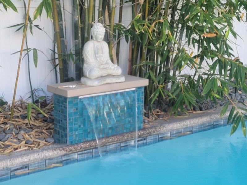 A serene Buddha statue sits atop a blue tiled water feature cascading into a swimming pool, framed by bamboo.