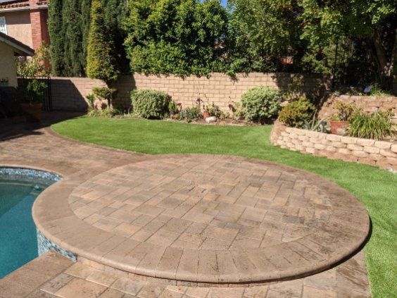 A circular stone patio next to a swimming pool, with a lush green lawn and a block wall in the background.