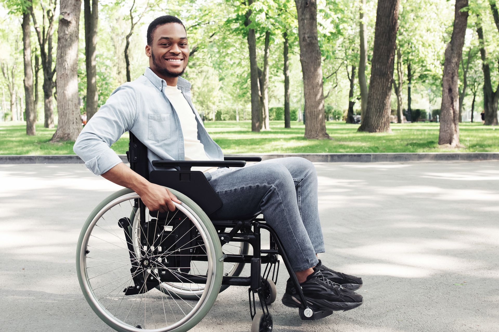 A smiling person in a wheelchair sits on a paved path in a sunlit park with trees in the background.