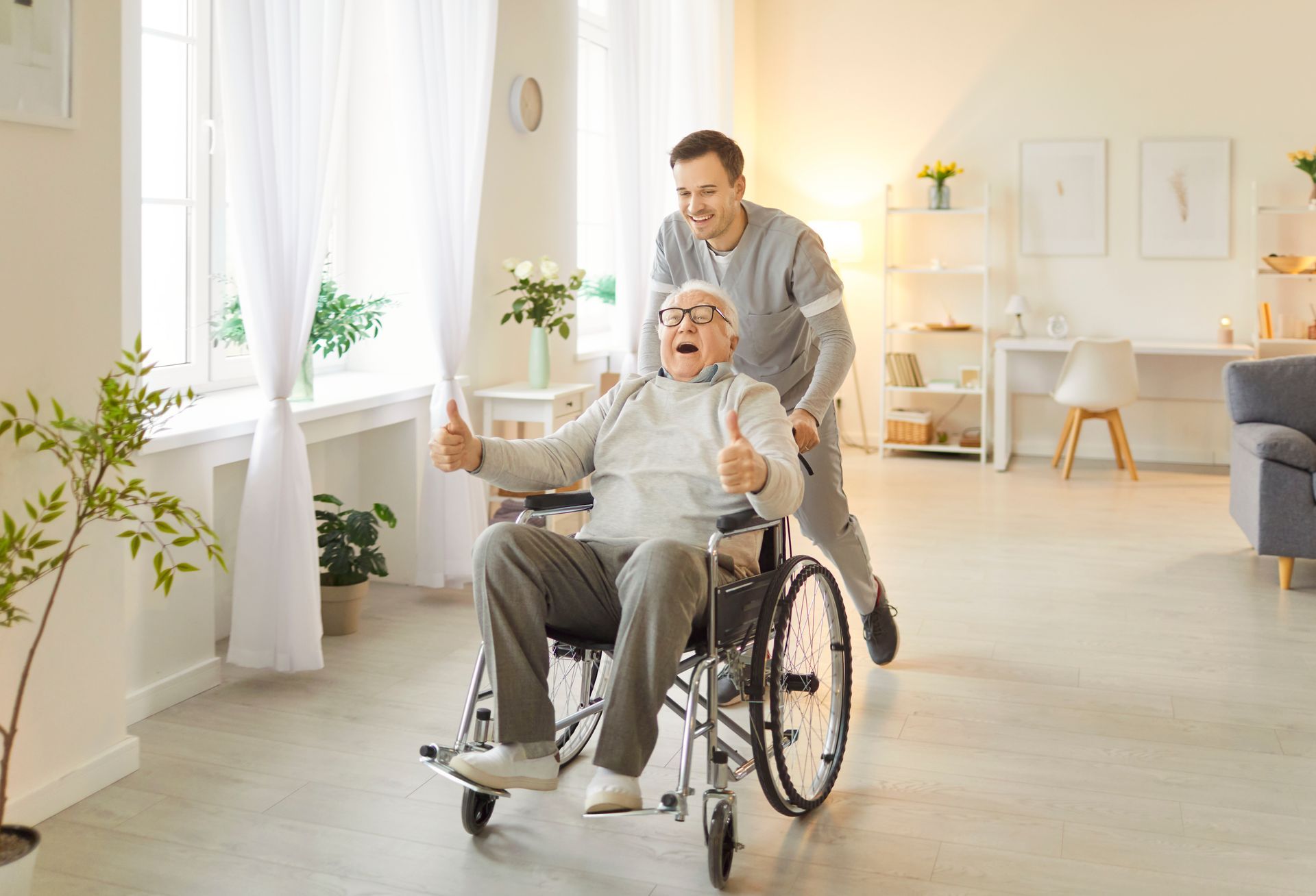 A person in a wheelchair gives a thumbs-up while being pushed by a caregiver in a bright, modern living room.
