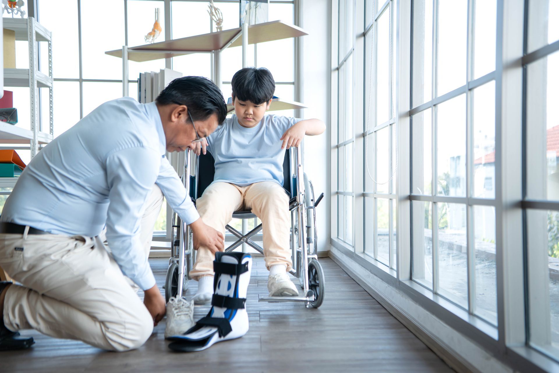 A caregiver kneeling to adjust a medical boot on a child sitting in a wheelchair indoors near large windows.