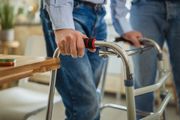 Hands grip the handles of a metal walker while standing, with a second person assisting nearby in a home setting.