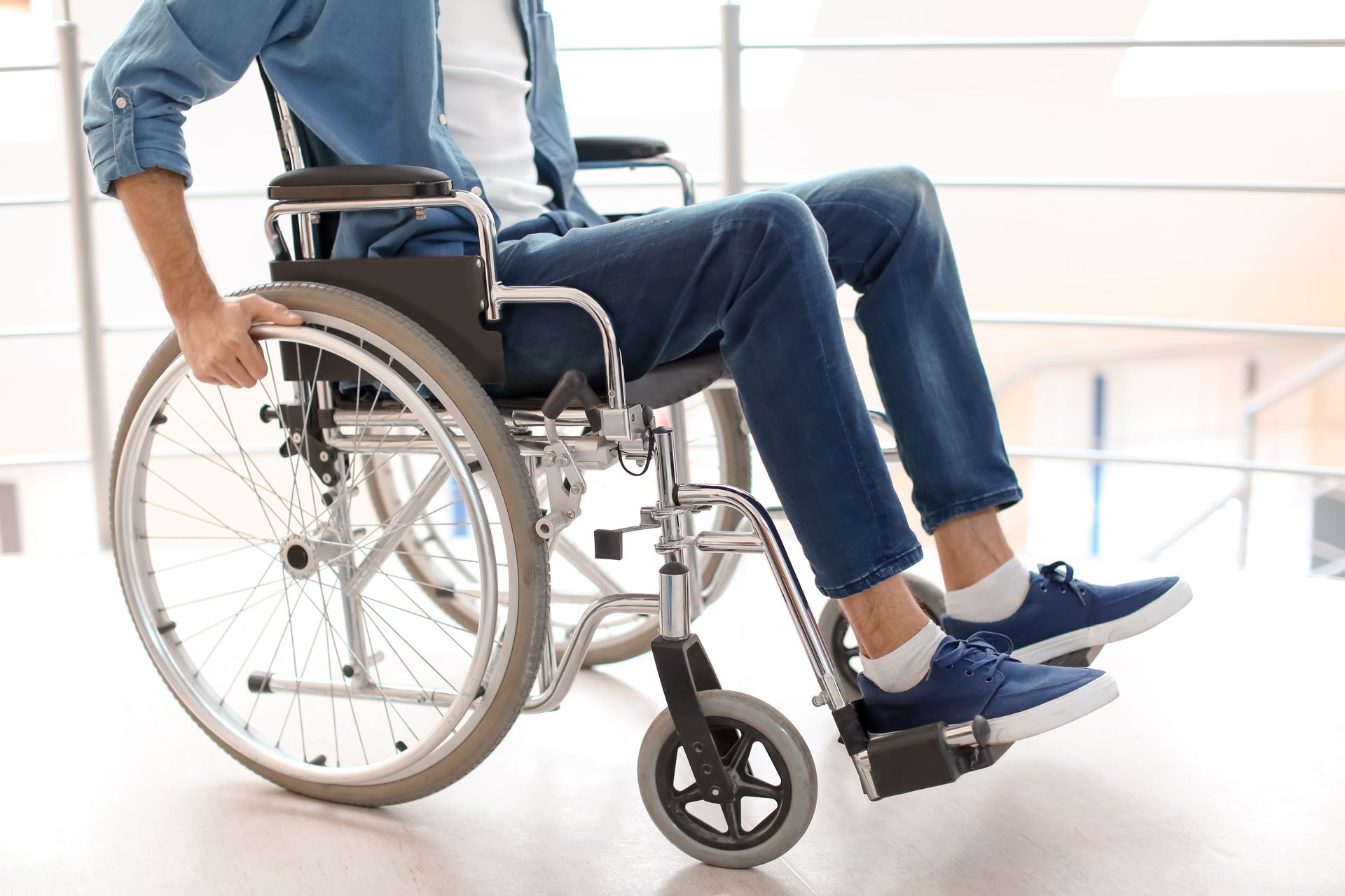 A person in jeans sits in a manual wheelchair indoors, with their hands on the wheel rims.
