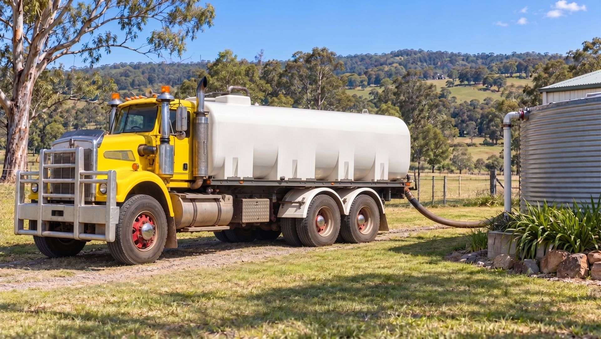 Yellow Water Tanker Truck Parked on Grass Beside a Farmhouse and Silo in a Rural Setting — Taree & Mid Coast Water Cartage in Forster, NSW