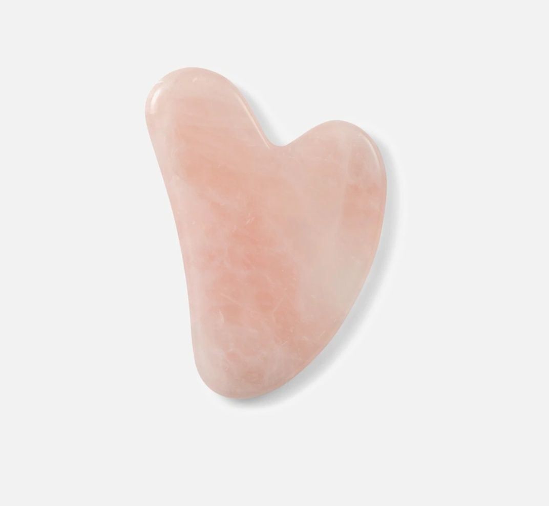Roze hartvormig gua sha instrument, gebruikt voor gezichtsmassage, tegen een witte achtergrond.